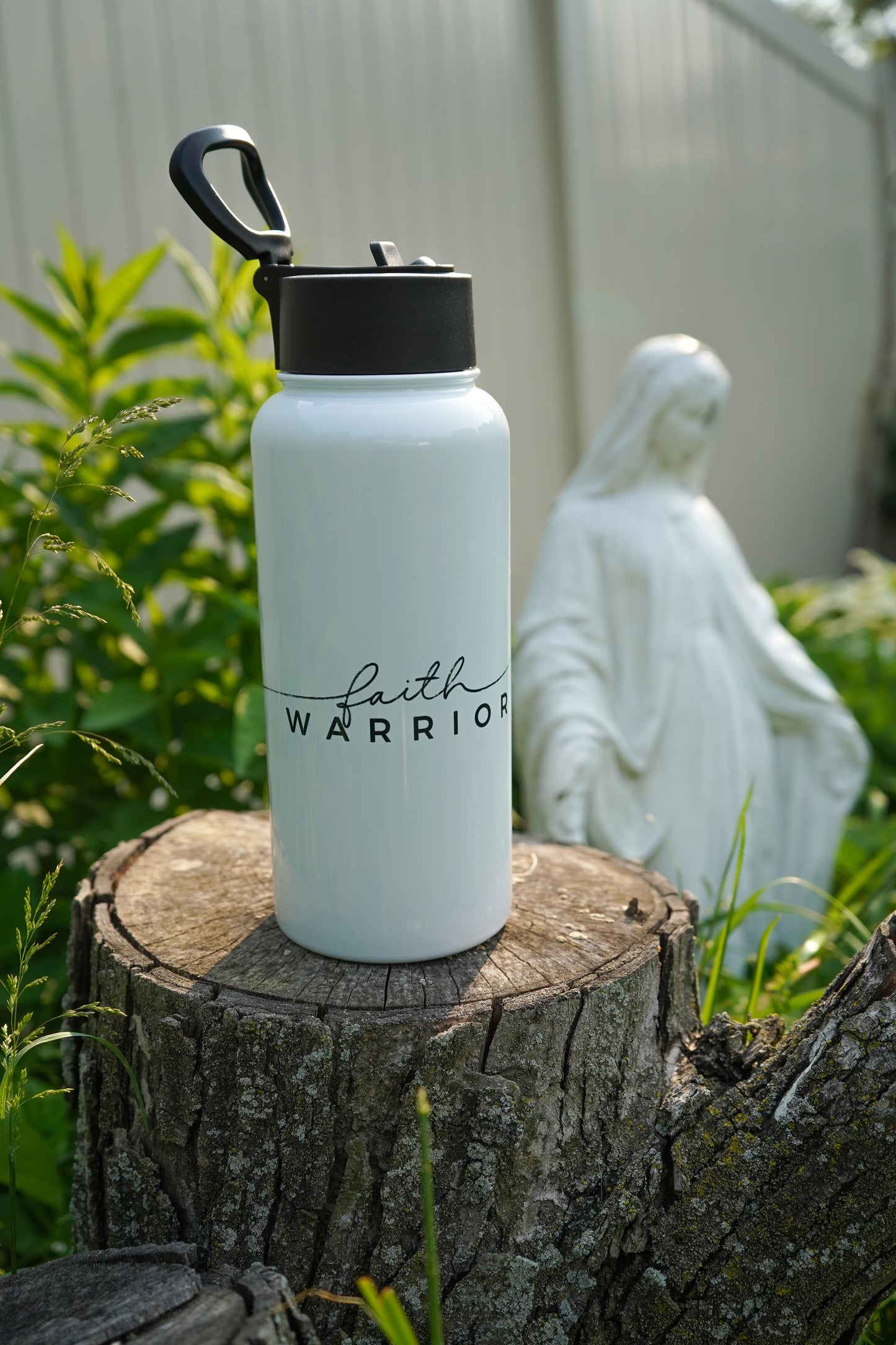 White water bottle with 'Faith Warrior' text on a tree stump with a statue in the background